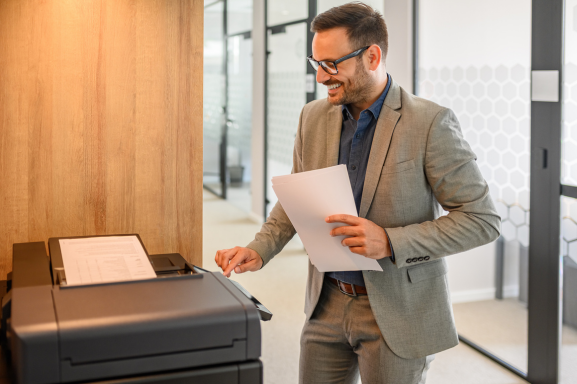 A cheerful professional in a modern office using a multifunction printer to complete a task. The individual holds printed documents while interacting with the device, highlighting the efficiency and versatility of multifunction printers in a business setting.